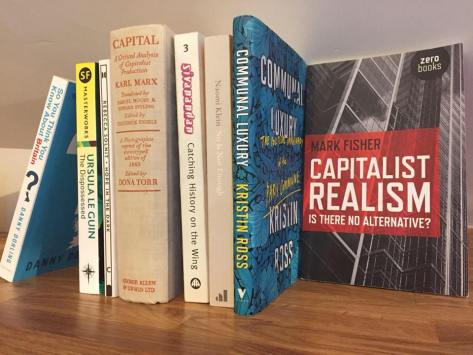 Picture of several books on a shelf, with Capitalist Realism by Mark Fisher, Capital by Karl Marx, Communal Luxury by Kristin Ross, and No is Not Enough by Naomi Klein clearly visible